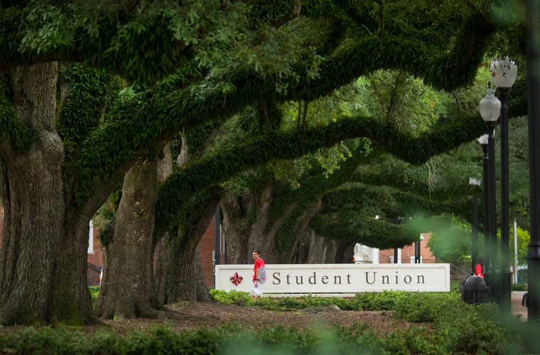 Student walking in front of the University of Louisiana at 69ý Student Union sign under a canopy of oak tree branches